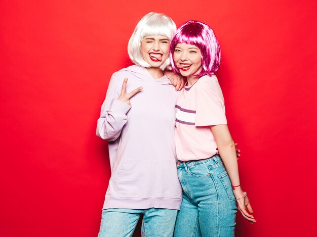 Two young sexy hipster girls in  wigs and red lips.Beautiful trendy women in summer clothes.Carefree models posing near red wall in studio going crazy.Shows tongue