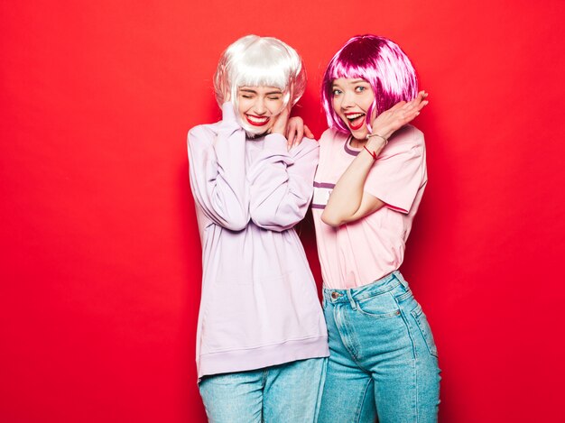 Two young sexy hipster girls in  wigs and red lips.Beautiful trendy women in summer clothes.Carefree models posing near red wall in studio going crazy