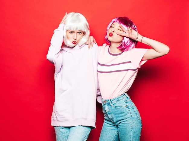 Free photo two young sexy smiling hipster girls in white wigs and red lips.beautiful trendy women in summer clothes.carefree models posing near red wall in studio going crazy