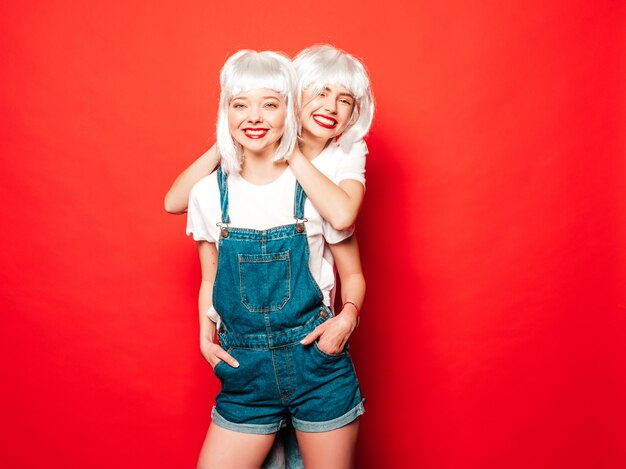 Две молодые сексуальные улыбающиеся хипстерские девушки в белых париках и красных губах. Красивые модные женщины в летней одежде. Беззаботные модели, позирует возле красной стены в студии летом сходит с ума