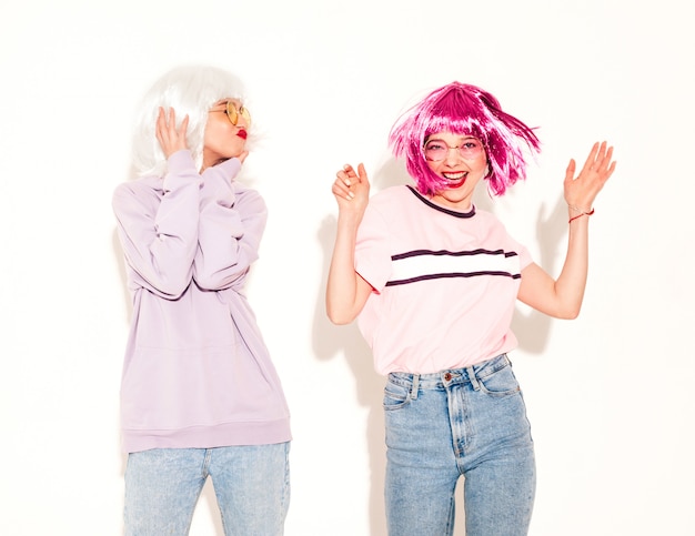 Two young sexy smiling hipster girls in wigs and red lips.Beautiful trendy women in  clothes.Carefree models posing near white wall in studio going crazy and dancing