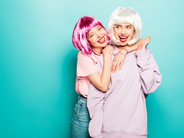 Two young sexy smiling hipster girls in wigs and red lips.Beautiful trendy women in summer clothes.Carefree models posing near blue wall in studio going crazy and hugging