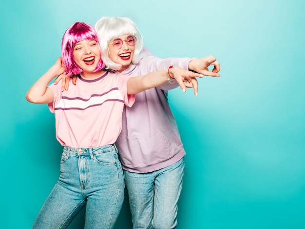 Two young sexy smiling hipster girls in wigs and red lips.Beautiful trendy women in summer clothes.Carefree models posing near blue wall in studio pointing on shop sales