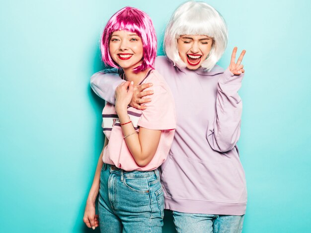 Two young sexy smiling hipster girls in wigs and red lips.Beautiful trendy women in summer clothes.Carefree models posing near blue wall in studio show peace sign