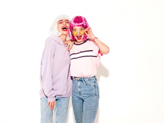 Two young sexy smiling hipster girls in wigs and red lips.Beautiful trendy women in summer clothes.Carefree models posing near white wall in studio going crazy and hugging