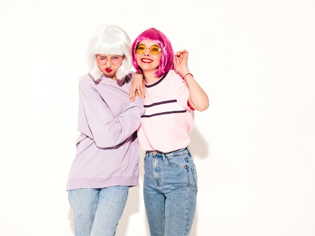 Two young sexy smiling hipster girls in wigs and red lips.Beautiful trendy women in summer clothes.Carefree models posing near white wall in studio going crazy and hugging
