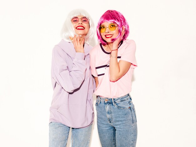 Two young sexy smiling hipster girls in wigs and red lips.Beautiful trendy women in summer clothes.Carefree models posing near white wall in studio going crazy and hugging