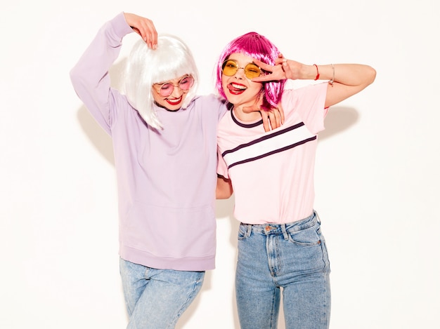 Two young sexy smiling hipster girls in wigs and red lips.Beautiful trendy women in summer clothes.Carefree models posing near white wall in studio shows peace sign