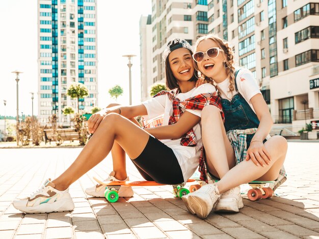 カラフルなペニースケートボードと2人の若い笑顔の美しい女の子。通りの背景に座っている夏の流行に敏感な服の女性。楽しくてクレイジーになるポジティブモデル