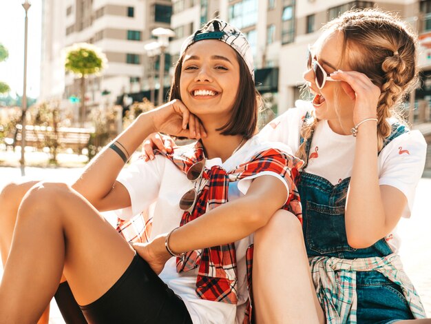 カラフルなペニースケートボードと2人の若い笑顔の美しい女の子。通りの背景に座っている夏の流行に敏感な服の女性。楽しくてクレイジーになるポジティブモデル