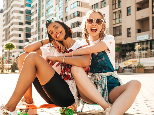 Two young smiling beautiful girls with colorful penny skateboards. Women in summer hipster clothes sitting in the street background. Positive models having fun and going crazy