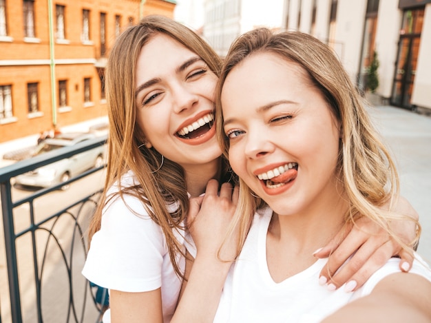 Two young smiling hipster blond women in summer white t-shirt clothes. Girls taking selfie self portrait photos on smartphone.  .Female showing tongue