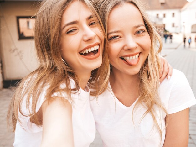 여름 흰색 티셔츠 옷에 두 젊은 미소 hipster 금발 여자. 스마트 폰에서 셀카 자기 초상화 사진을 찍는 여자. 거리 배경에 포즈 모델.