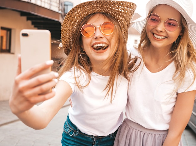 Two young smiling hipster blond women in summer white t-shirt clothes. Girls taking selfie self portrait photos on smartphone. 