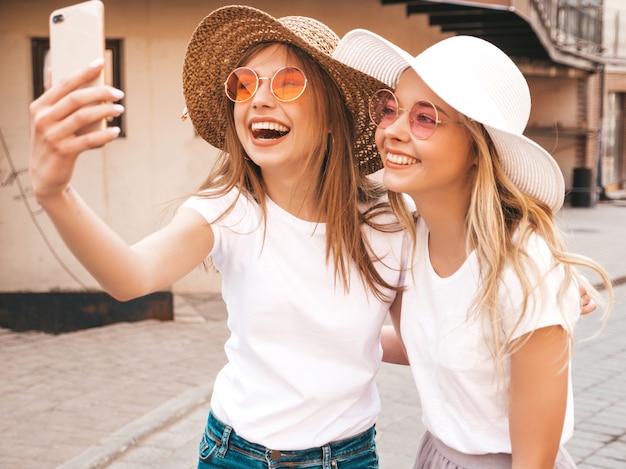 Two young smiling hipster blond women in summer white t-shirt clothes. Girls taking selfie self portrait photos on smartphone. 