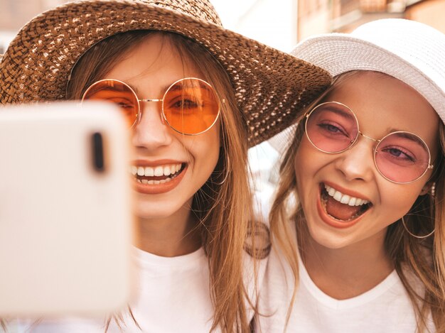 Two young smiling hipster blond women in summer white t-shirt clothes. Girls taking selfie self portrait photos on smartphone. 