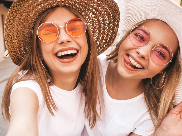 Two young smiling hipster blond women in summer white t-shirt clothes. Girls taking selfie self portrait photos on smartphone. 