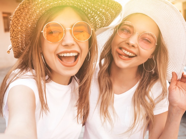 Foto gratuita due giovani donne bionde sorridenti dei pantaloni a vita bassa in vestiti bianchi della maglietta di estate. ragazze che prendono le foto dell'autoritratto del selfie sullo smartphone.
