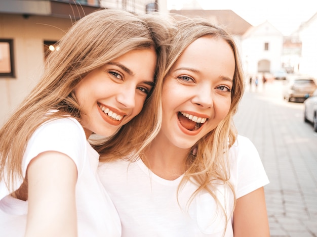 Two young smiling hipster blond women in summer white t-shirt clothes. Girls taking selfie self portrait photos on smartphone. 