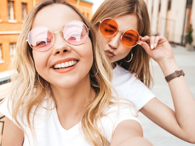 夏に2人の若い笑顔ヒップスターブロンド女性白いtシャツ服。スマートフォンでセルフポートレート写真を撮る女の子。