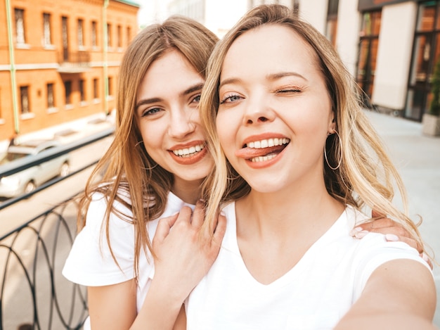 Free photo two young smiling hipster blond women in summer white t-shirt clothes. girls taking selfie self portrait photos on smartphone.  .