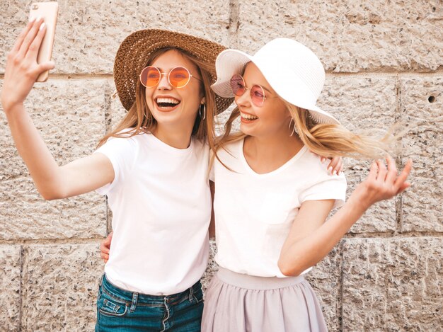 Two young smiling hipster blond women in summer white t-shirt. Girls taking selfie self portrait photos on smartphone.Models posing on street background.Female shows positive emotions