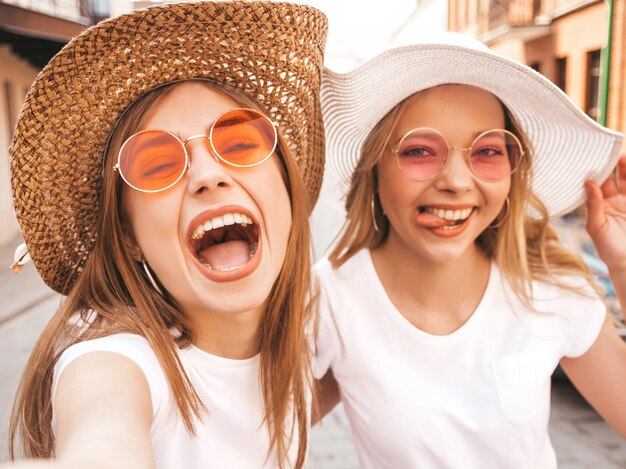 夏に2人の若い笑顔ヒップスターブロンド女性白いtシャツ。スマートフォンでセルフポートレート写真を撮る女の子。通りの背景でポーズをとるモデル。女性は肯定的な感情を示しています。