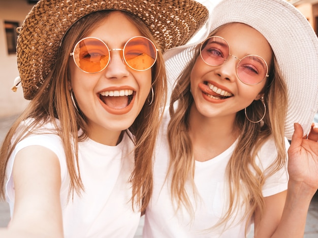 Two young smiling hipster blond women in summer white t-shirt. Girls taking selfie self portrait photos on smartphone.Models posing on street background.Female shows tongue and positive emotions