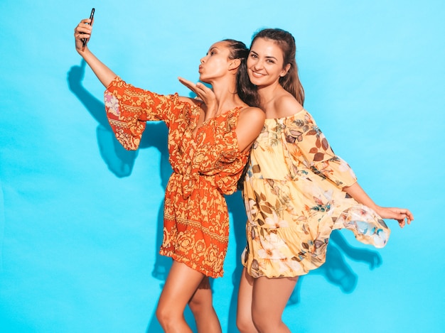 여름 히피 드레스에 두 젊은 미소 hipster 여성. 스마트 폰에 셀카 자기 초상화 사진을 찍는 소녀. 스튜디오에서 파란색 벽 근처 포즈 모델. 여성 공기 키스를 제공합니다