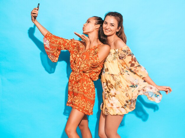 Two young smiling hipster women in summer hippie dresses.Girls taking selfie self portrait photos on smartphone.Models posing near blue wall in studio.Female gives air kiss