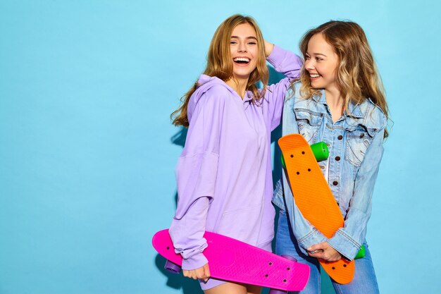 Две молодые стильные улыбающиеся белокурые женщины с пенни скейтбордами. Модели в летних битник спортивной одежды позирует возле синей стены. Позитивные женщины сходят с ума