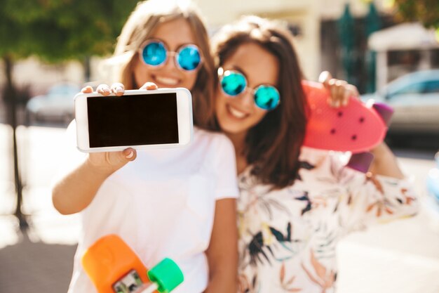 Две молодые стильные улыбающиеся хиппи брюнетки в летний солнечный день в хипстерских одеждах с пенни скейтбордом позируют