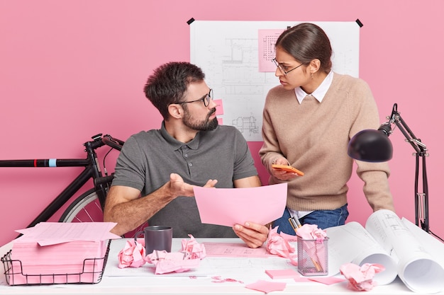 Two young woman and man coworkers look angrily at each other blame for mistake pose at modern office desk discuss sketch for building project. Professional engineers collaborate on blueprints