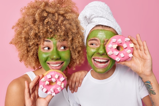 두 젊은 여성이 녹색 보습 마스크를 바르고 마시멜로가 있는 달콤한 도넛을 들고 분홍색 배경에 격리된 캐주얼한 티셔츠를 입고 매우 행복합니다. 여성 모델은 미용 치료를 받습니다