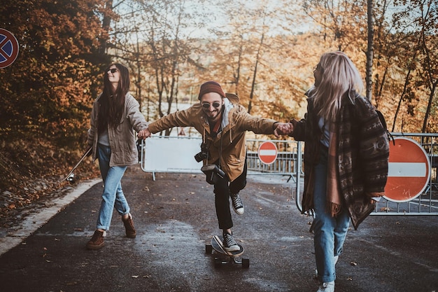 두 젊은 여성이 가을 공원을 산책하면서 친구에게 스케이트보드 타는 법을 가르치고 있습니다.