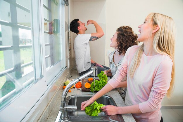 Two young women cleaning the lettuce vegetable laughing while looking at man eating carrot