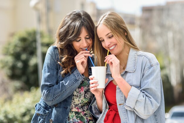 同じ飲み物をしている2人の若い女性が屋外で2つのストローと一緒にガラスを奪う。