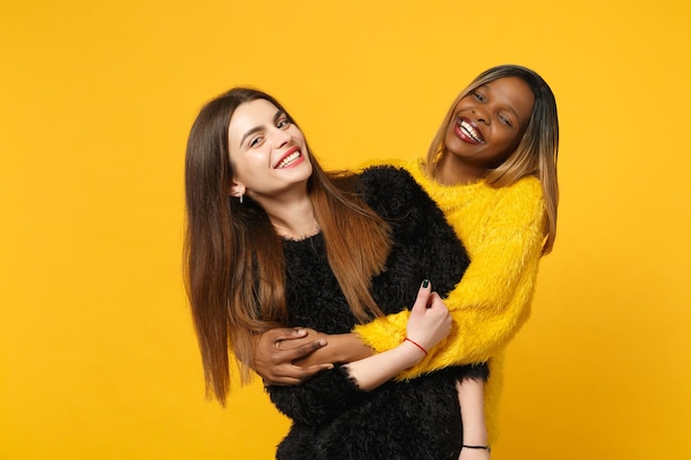 Two young women friends european and african american in black yellow clothes standing posing isolated on bright orange wall background, studio portrait. people lifestyle concept. mock up copy space.