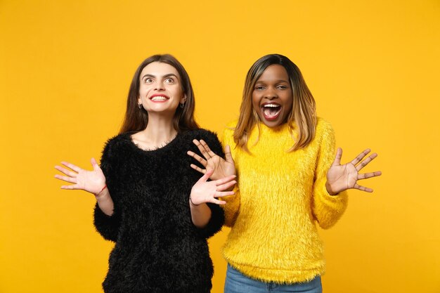 明るい​オレンジ色​の​壁​の​背景​、​スタジオ​の​肖像画​に​分離された​ポーズ​で​立っている​黒​黄色​の​服​を​着た​ヨーロッパ人​と​アフリカ系​アメリカ人​の​2​人​の​若い​女性​の​友人​。​人々​の​ライフスタイル​の​コンセプト​。​コピー​スペース​を​モックアップ​します​。