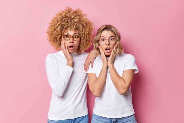 Two young women gasp in surprise stare shocked dessed in casual clothes isolated over pink background Amazed ladies hear exciting news Emotions reactions and human face expressions concept