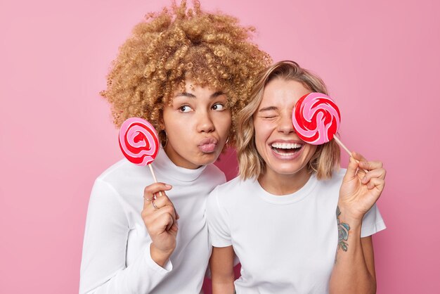 2人の若い女性は、白い服を着たロリポップで楽しいポーズをとっています。愚かな周りはピンクの背景の上に甘い歯を分離しています幸せな友達はスティックにキャラメルキャンディーを持っています屋内