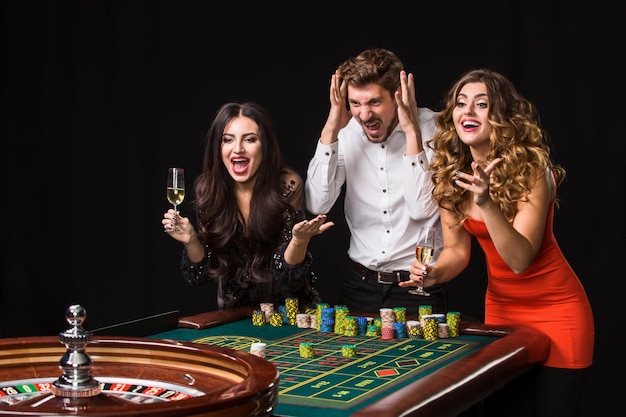 Two young women and man behind roulette table on black background. Emotions players