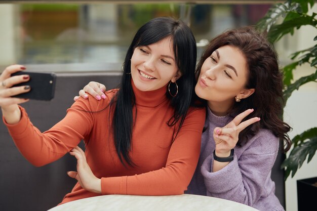 Two young women taking selfie in cafe while sitting at table, smiling and showing v sign, friends spending time together.