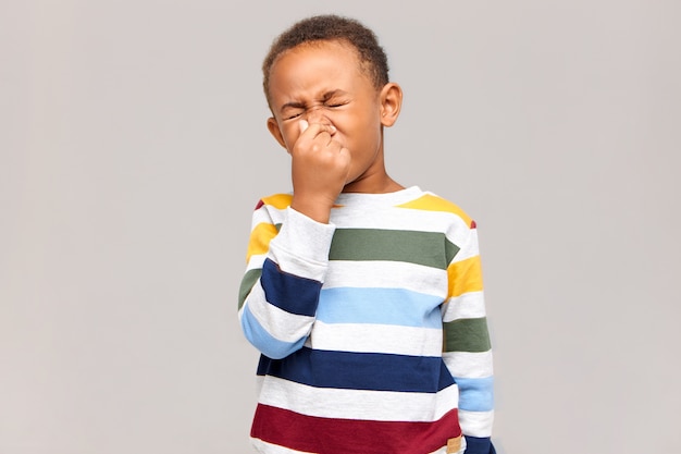 うーん、嫌な！悪臭や悪臭のために目を閉じて鼻をつまんでいる感情的な嫌悪感のあるアフリカ系アメリカ人の少年の肖像画。アレルギー、くしゃみをしている浅黒い肌の男児