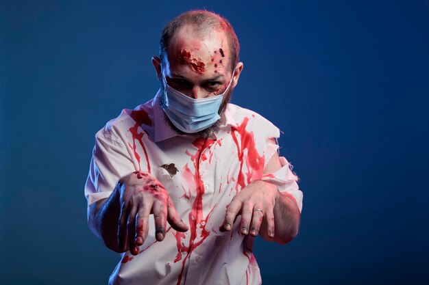 코비드 19 얼굴 마스크를 쓰고 피 묻은 상처와 공격적인 표정을 가진 못생긴 할로윈 좀비. 무서운 악마의 눈과 무서운 얼굴을 가진 소름 끼치는 죽은 시체, 전염병 동안 뇌를 먹고 있습니다.