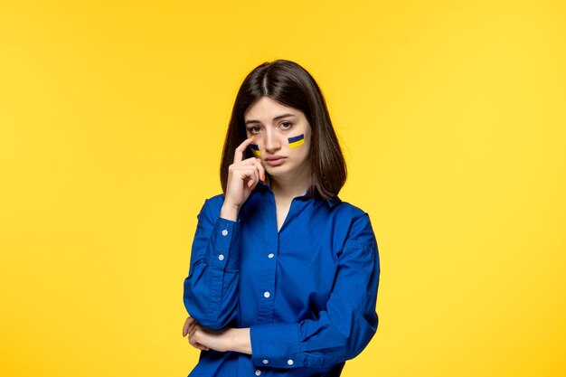Ukraine russian conflict cute young girl in blue shirt yellow background wiping tear off cheek