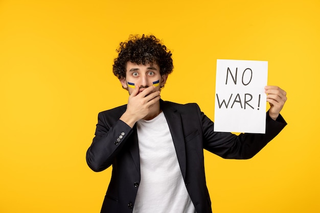 Free photo ukraine russian conflict young guy in black blazer with ukrainian flag on face holding no war sign