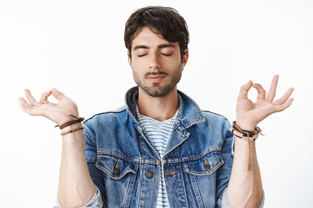 Free photo unaltered shot of good-looking mature male with beard and wavy dark hair standing with peaceful expression and closed eyes in lotus pose with zen gesture, meditating keeping calm over gray wall