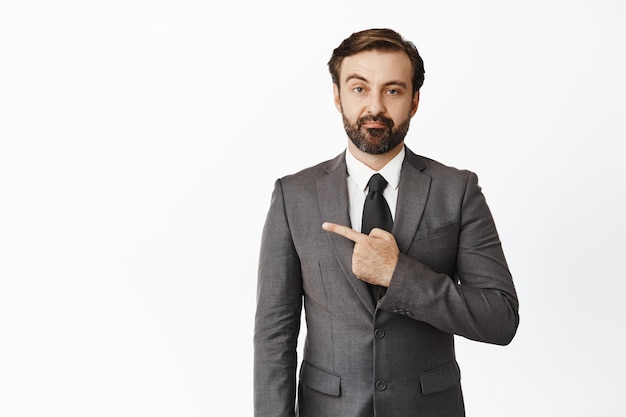 Unamused bearded male entrepreneur wearing suit pointing left and looking with disbelief standing against white background