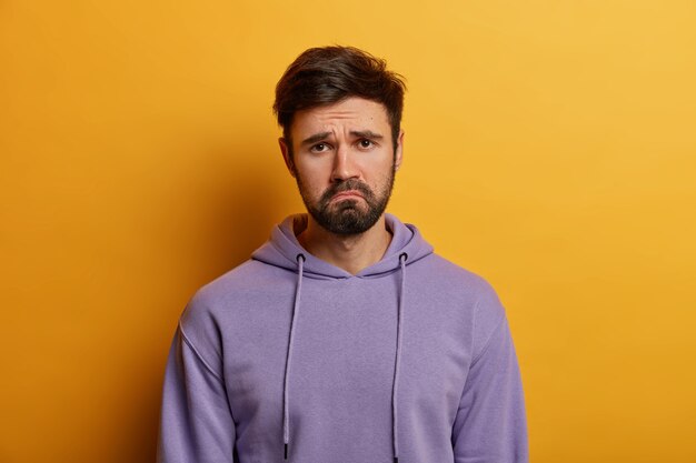 Unamused sad miserable guy purses lips, frowns face with dissatisfaction, wears hoodie, poses indoor against yellow wall, sulks troubled as someone hurt his feelings, wears purple hoodie